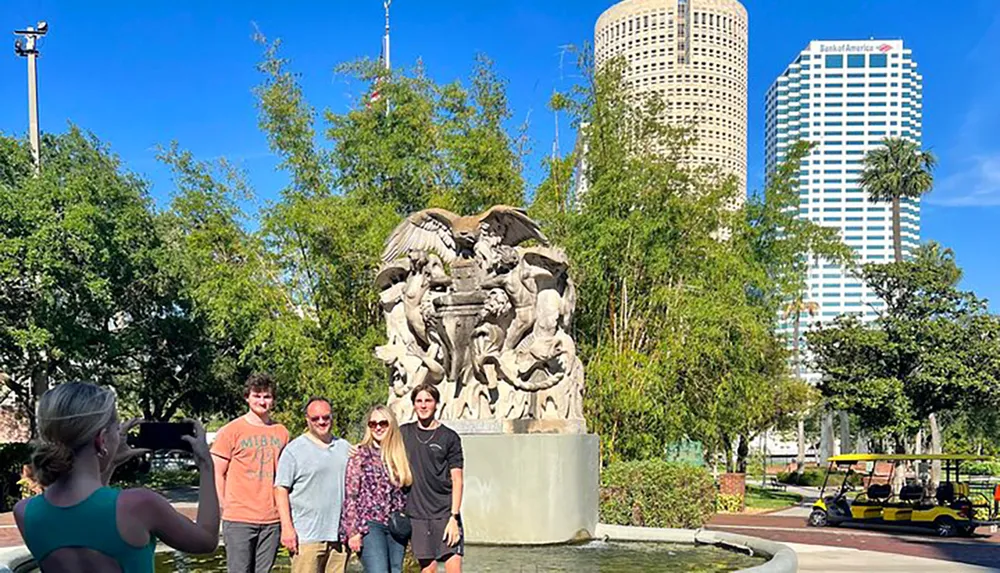 A group of people poses for a photo in front of a large sculpture in a park with tall buildings in the background