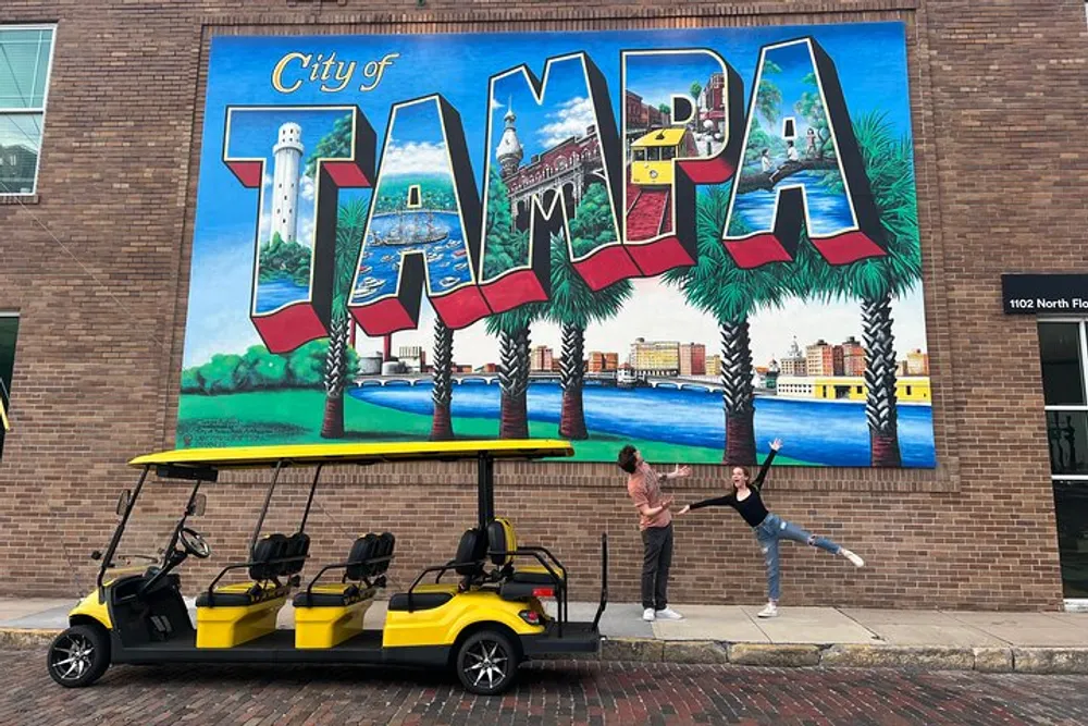 Two people are playfully interacting in front of a colorful mural that reads City of Tampa with a parked yellow golf cart in the foreground
