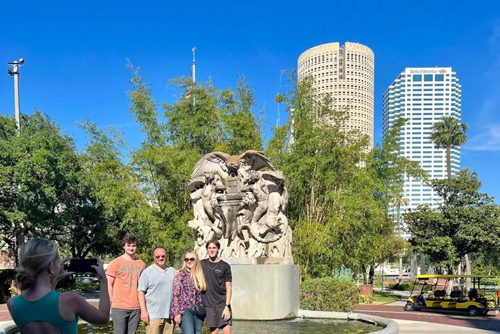 A group of people is posing for a photo in front of an intricate sculpture with city skyscrapers in the background on a sunny day