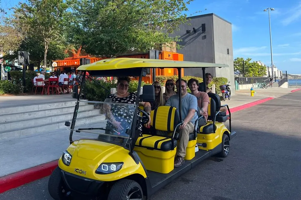 A group of people is smiling for a photo while sitting in a yellow golf cart on a sunny day with an outdoor seating area visible in the background