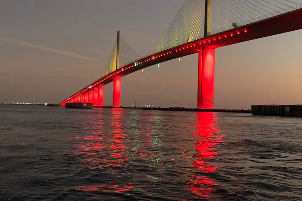 A long cable-stayed bridge illuminated with red lights is reflected in the water below during twilight
