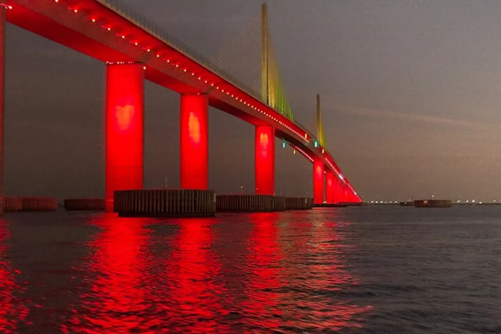 The image shows a long bridge illuminated with red lighting over a body of water reflecting a striking red hue on the surface