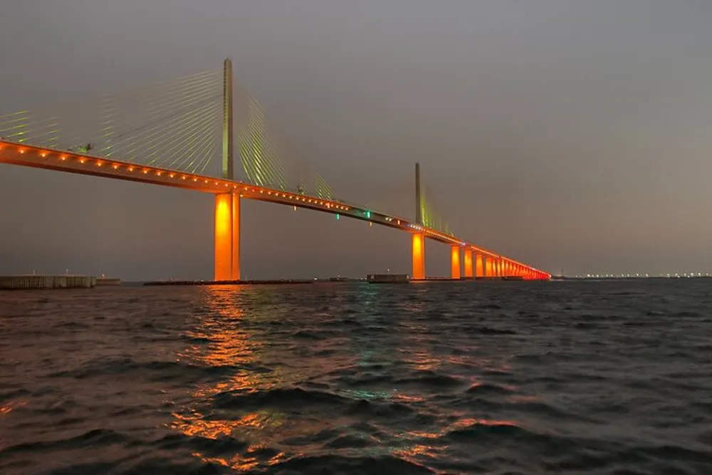 The image shows an illuminated suspension bridge spanning across a body of water under a dusk or dawn sky