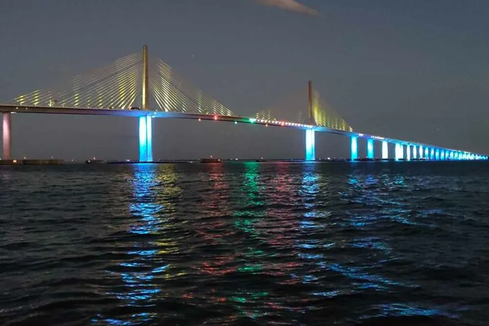 The image shows a long bridge at night illuminated with colorful lights reflecting on the surface of the water below