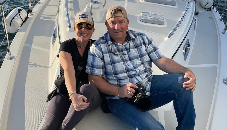 A man and a woman are smiling while seated on the deck of a boat, with the man holding a camera.