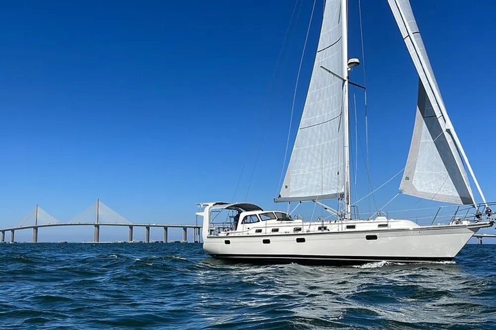 A sailboat is cruising on clear waters near a long bridge on a sunny day