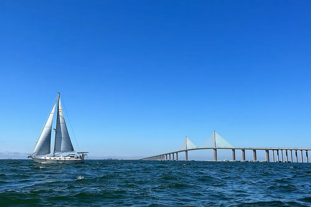 A sailboat is cruising on the water near a long bridge under a clear blue sky