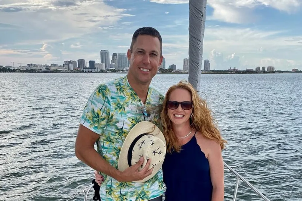 A smiling man and woman are posing together on a boat with a coastal city skyline in the background