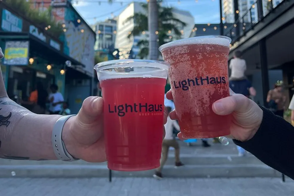 Two people are holding up plastic cups of red beverages with the LightHaus logo in what appears to be an outdoor social setting