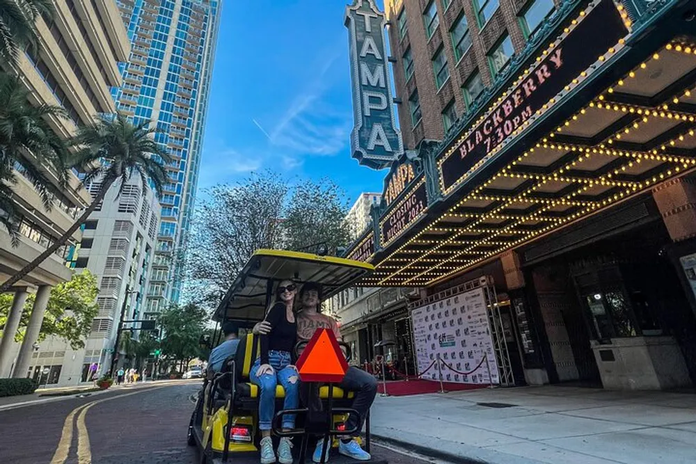 Two people are sitting in a yellow golf cart in front of the Tampa Theatre which has a brightly lit marquee and a red carpet event setup set against a backdrop of modern buildings under a clear sky