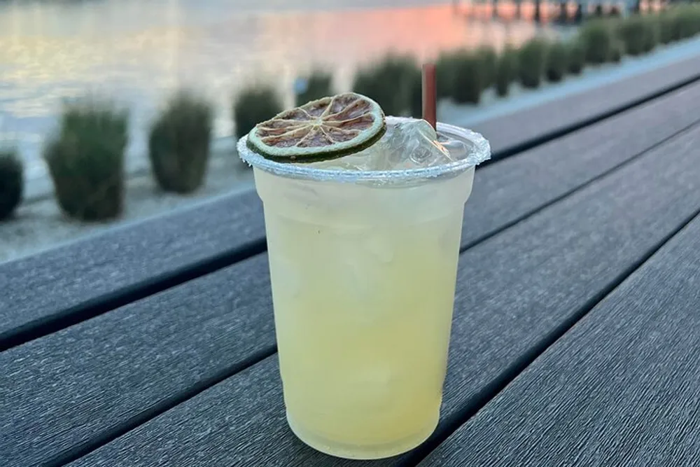 A refreshing cocktail with a lime garnish sits on a wooden table with a tranquil water view in the background