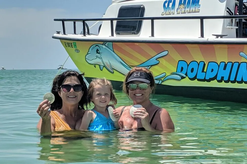 Three people are smiling and standing in clear shallow water near a boat that has DOLPHINS written on the side and they are holding seashells
