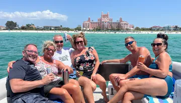 A group of five people are smiling and enjoying a sunny boat ride near a beach with a large pink building in the background.