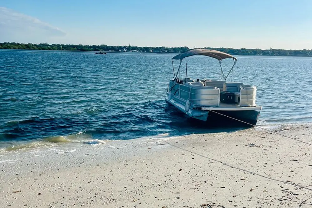 A pontoon boat is moored near a sandy shore with a view of the calm blue water and another boat in the distance