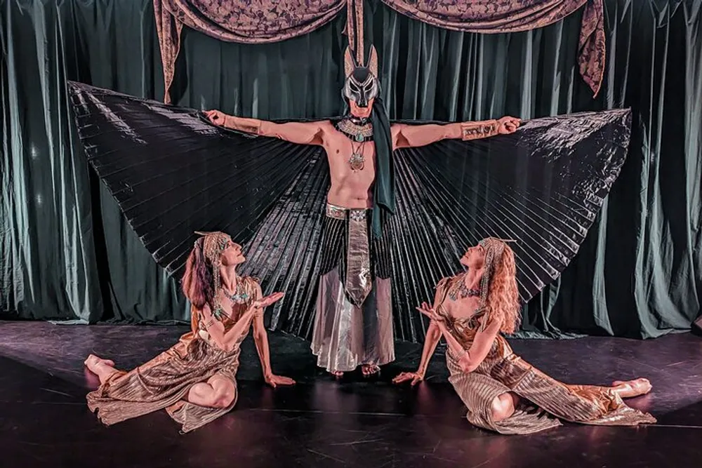 A person adorned with an elaborate avian costume spreads their wings while two others are seated at their sides gazing upwards against a backdrop of curtains on stage
