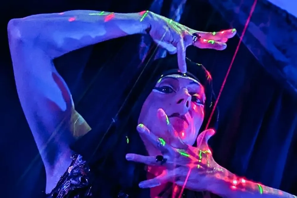 A person wearing dark clothing is dramatically posing with their arms raised illuminated by colorful spotlights against a dark background