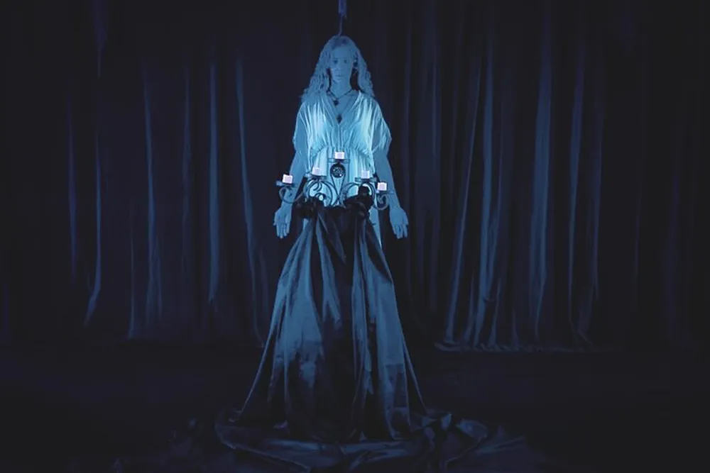 A person stands in a flowing costume illuminated by a blue light against a backdrop of dark curtains