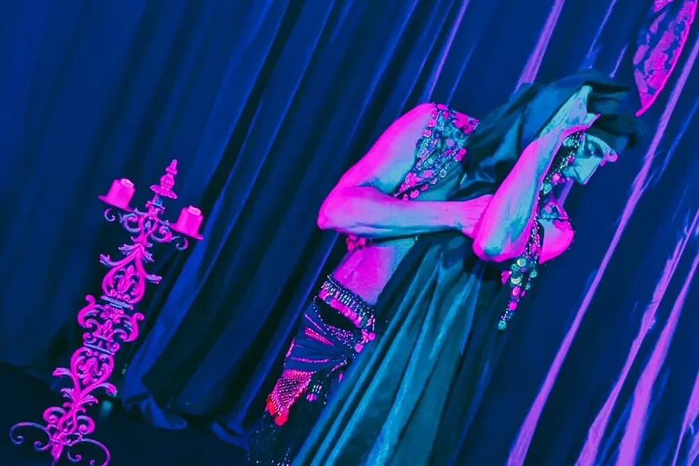 This image portrays an individual in a stylized costume posing next to a candelabra under vibrant pink and blue lighting