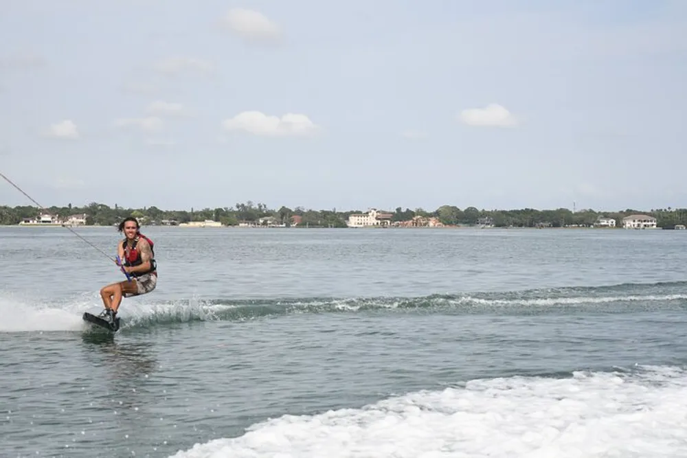 A person is wakeboarding on a body of water with a clear sky above and buildings in the distance