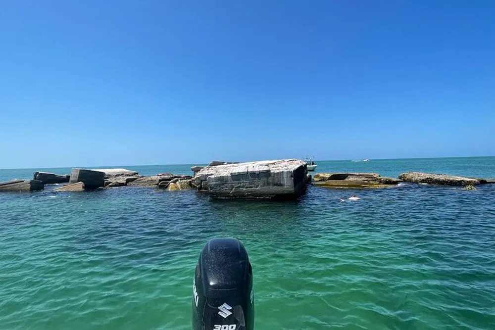 The image shows a view from a boat featuring sunken concrete structures partially submerged in clear shallow greenish-blue water set under a clear blue sky