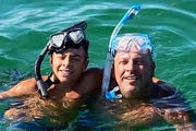 Two people, likely a child and an adult, are smiling at the camera while wearing snorkeling gear and floating in clear water.