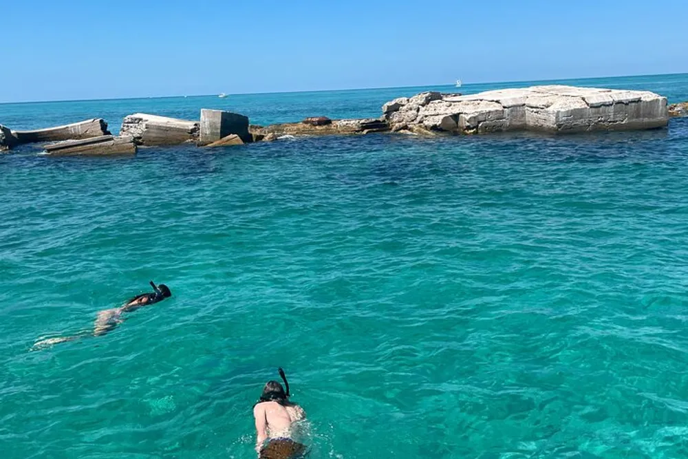 Two people are snorkeling in clear blue water near rocky ruins under a bright blue sky
