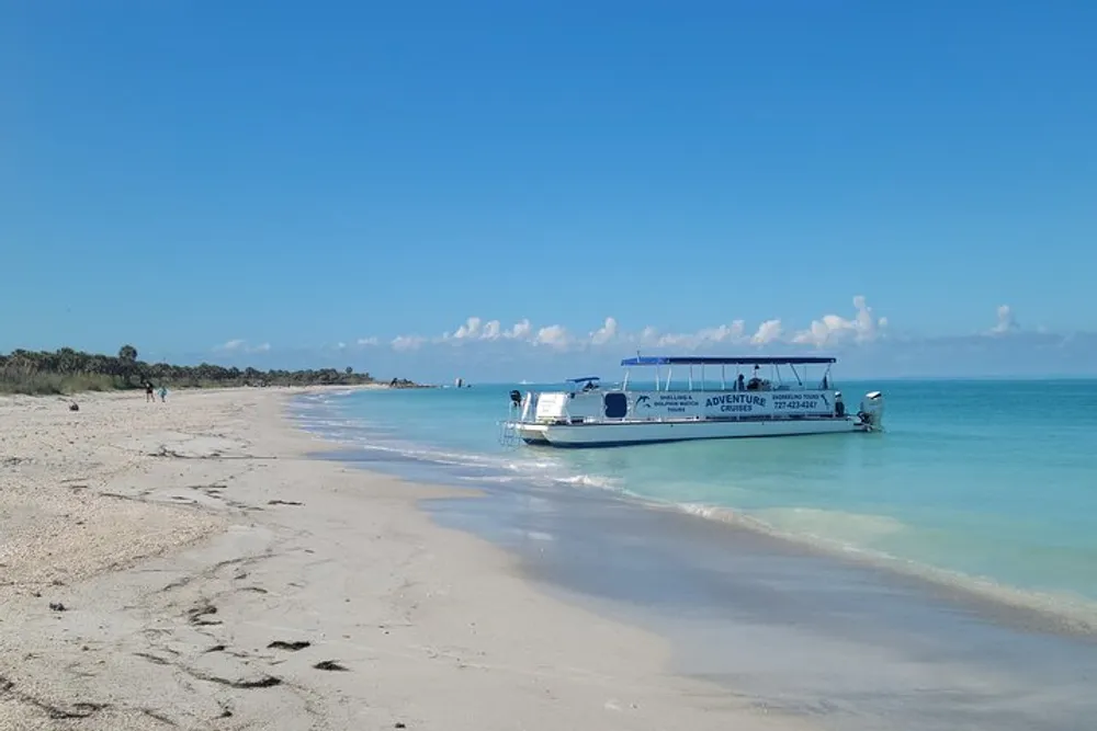 A boat is anchored near the shore of a beautiful beach with clear blue skies above and a couple of individuals visible in the distance walking along the sand