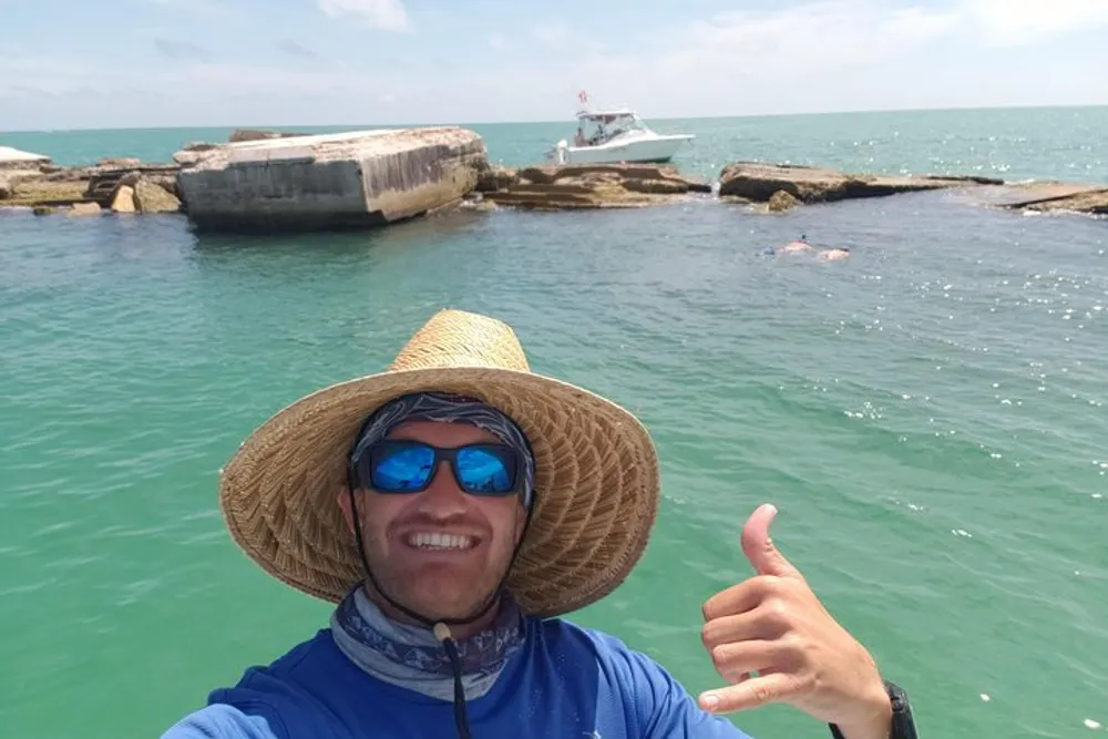 A person wearing a wide-brimmed hat and sunglasses is giving a thumbs up while taking a selfie with a boat and clear blue water in the background