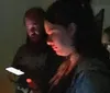 In a dimly lit room a person is looking intently at their smartphone screen illuminating their face while another person appears in the background with a faint glow on their face