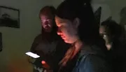 In a dimly lit room, a person is looking intently at their smartphone screen, illuminating their face, while another person appears in the background with a faint glow on their face.