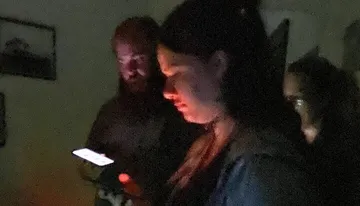 In a dimly lit room, a person is looking intently at their smartphone screen, illuminating their face, while another person appears in the background with a faint glow on their face.