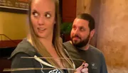 A woman holding divining rods looks to the side while a man standing behind her gazes at her.