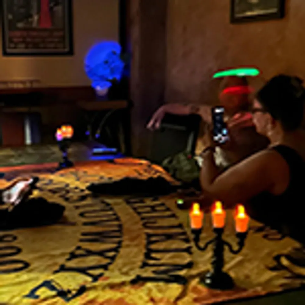 A woman appears surprised or amusingly shocked with wide eyes while a man looks on from behind with an amused expression in a dimly lit indoor setting