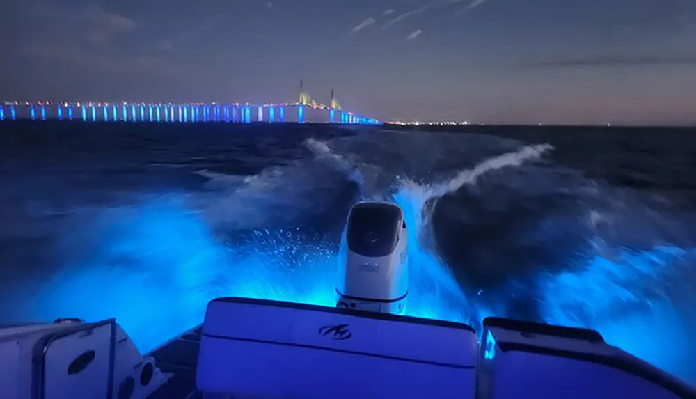 A boat creates a glowing blue wake at night with an illuminated bridge in the background