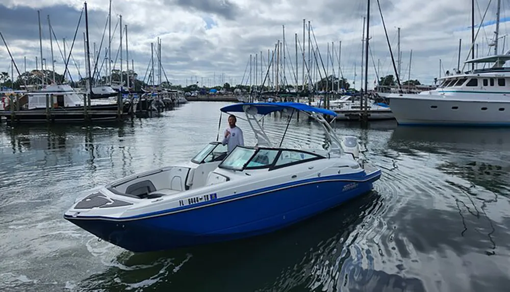 A person stands at the helm of a blue and white motorboat moving through a marina dotted with various docked boats under a cloudy sky