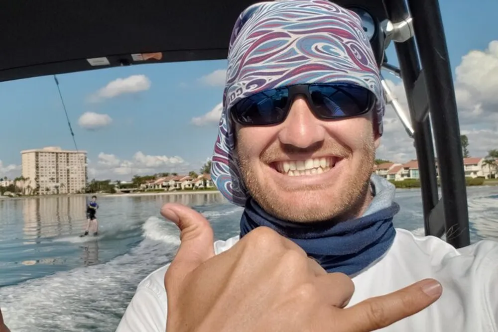 A person wearing sunglasses and a bandana gives a thumbs-up while someone is wakeboarding in the background