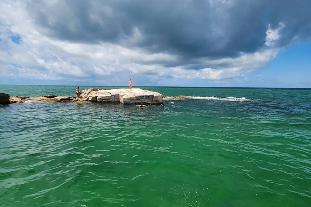People enjoy swimming near a concrete structure in a green-blue sea under a dramatic cloudy sky