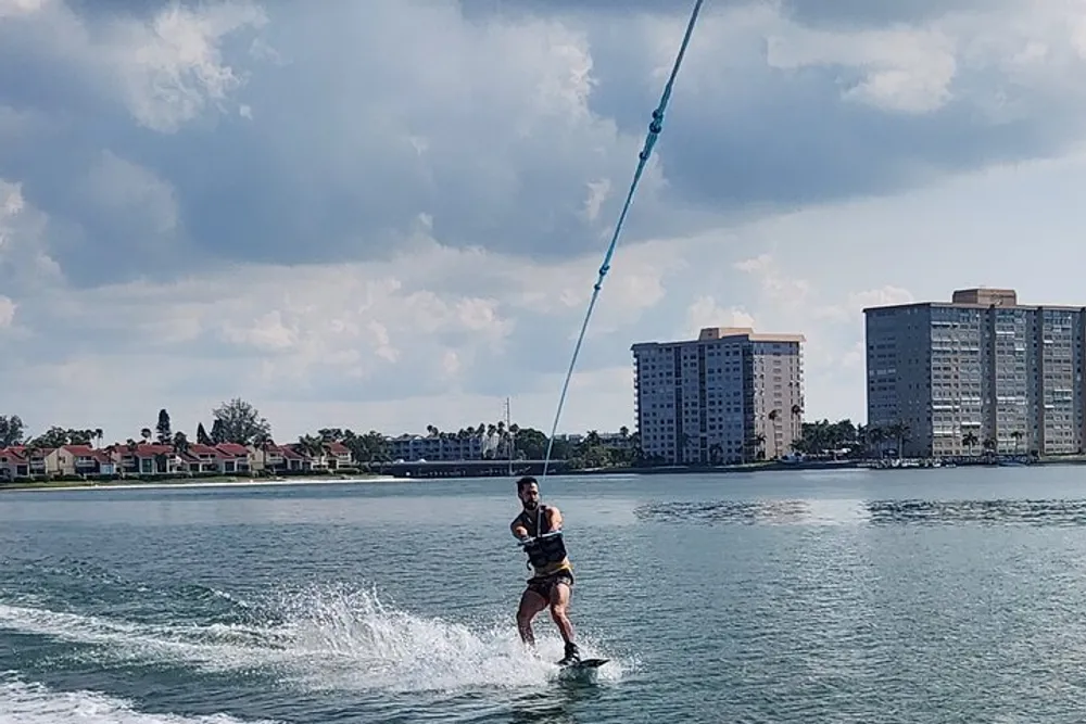 A person is wakeboarding on a body of water with a clear sky and coastal buildings in the background