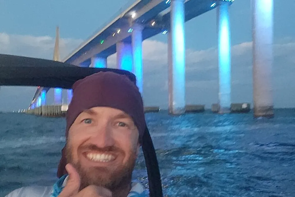 A man is smiling for a selfie with a lit-up bridge in the background during dusk or evening hours
