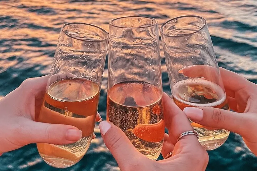 Three people are toasting with glasses of sparkling beverage over a water backdrop bathed in the warm glow of a sunset