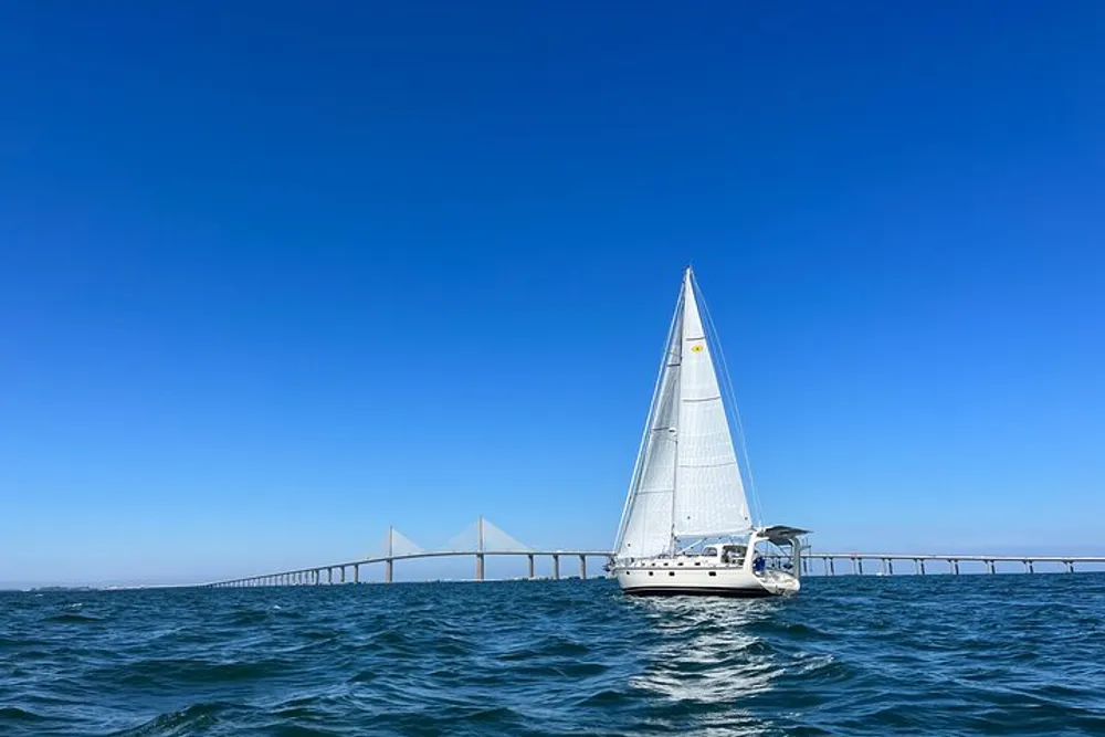 A sailboat is cruising on the water with a long bridge extending across the background under a clear blue sky