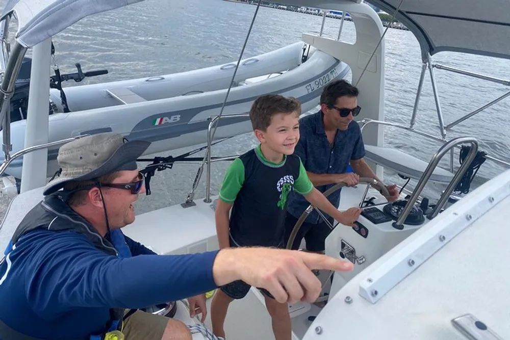 Three people one of whom is a child are on a sailboat with one man pointing at something out of frame and the child appears to be steering under the watchful eyes of the adults