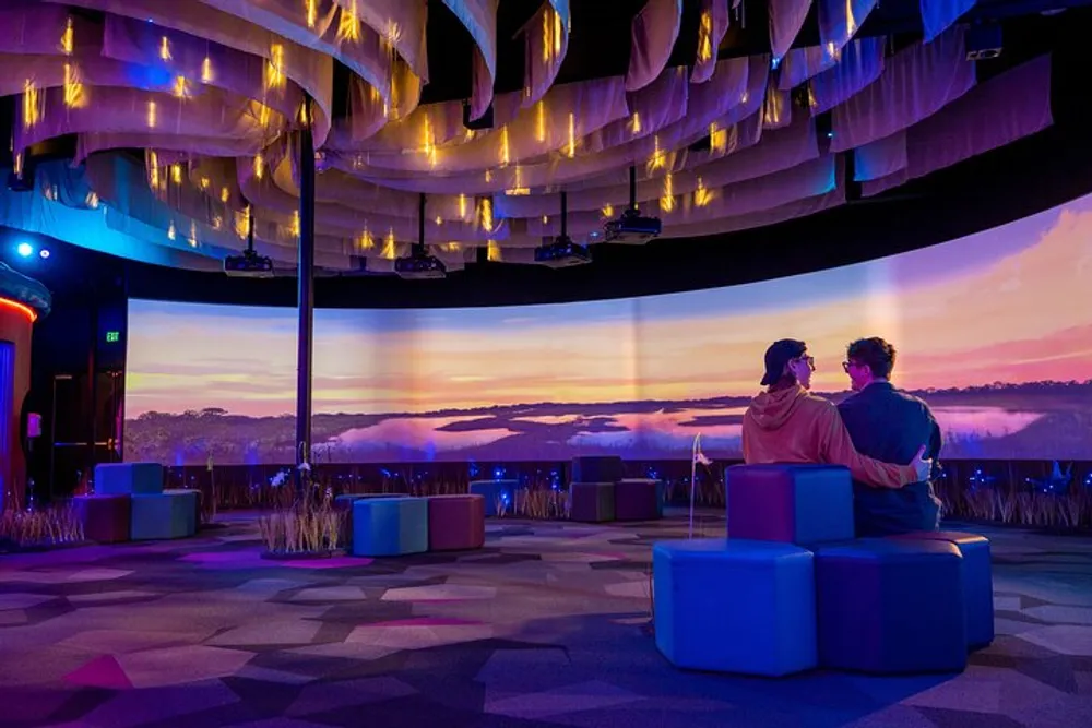 Two individuals are seated on colorful blocks enjoying a vibrant sunset scenery projected on a panoramic screen inside a modern room with artistic decor
