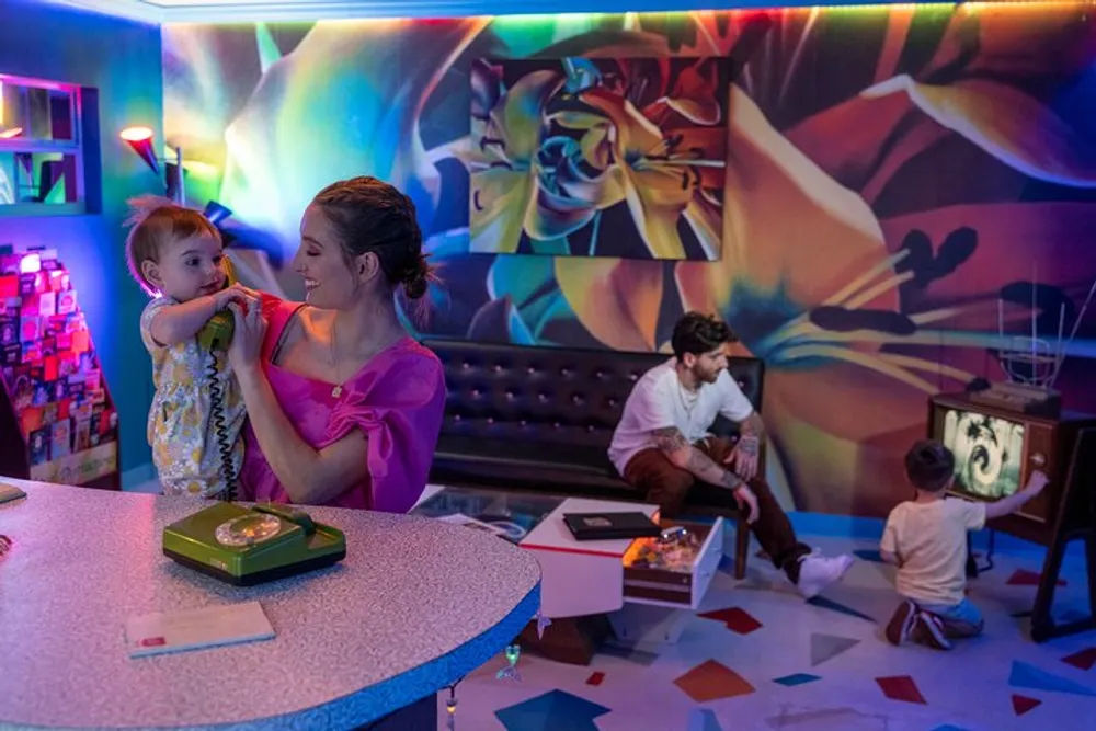 A family is spending time in a colorful room with vibrant murals where a woman is holding a young child and a man is watching another child play with a toy television
