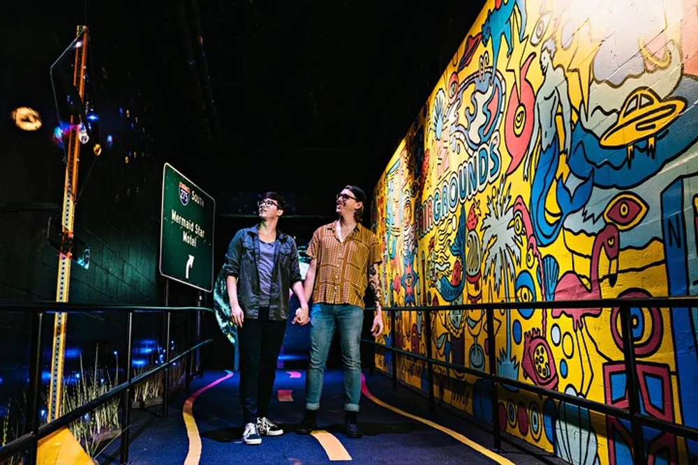 Two individuals are walking next to a vibrantly colored mural with amusement park signs indicating attractions in a dark gallery-like space