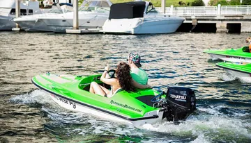 Two individuals are riding a green mini powerboat in a marina with other boats and yachts docked in the background.