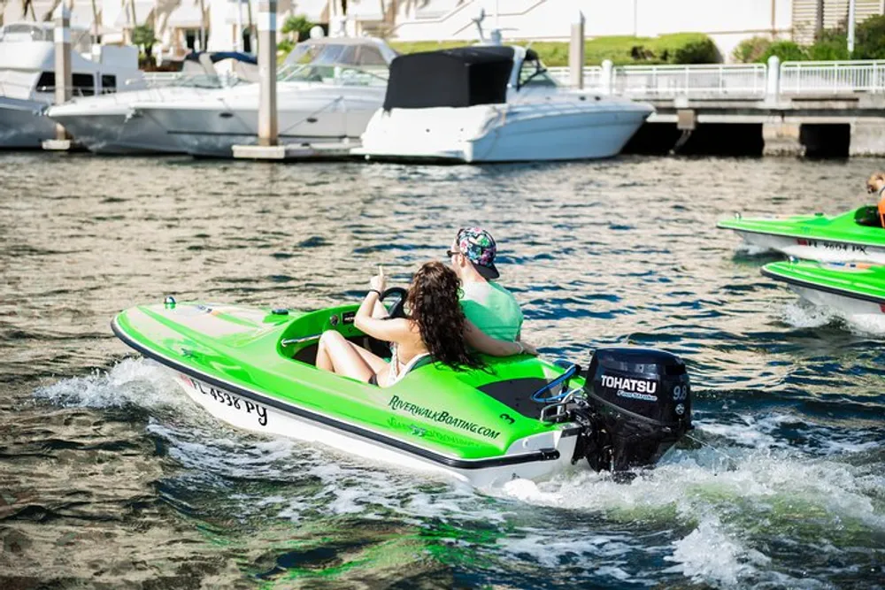 Two individuals are riding a green jet ski on the water near docked boats on a sunny day