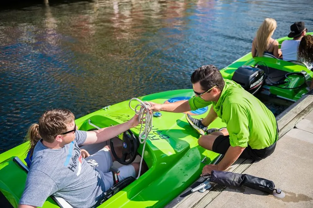 A person in a bright green shirt assists another individual in a lime-green boat as two others sit in a similar boat nearby docked on a sunny waterway