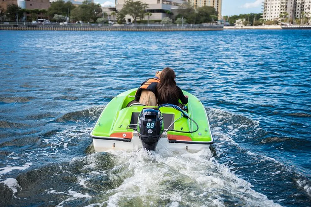 A person in a life jacket is operating a green and white personal watercraft on a body of water with buildings in the background