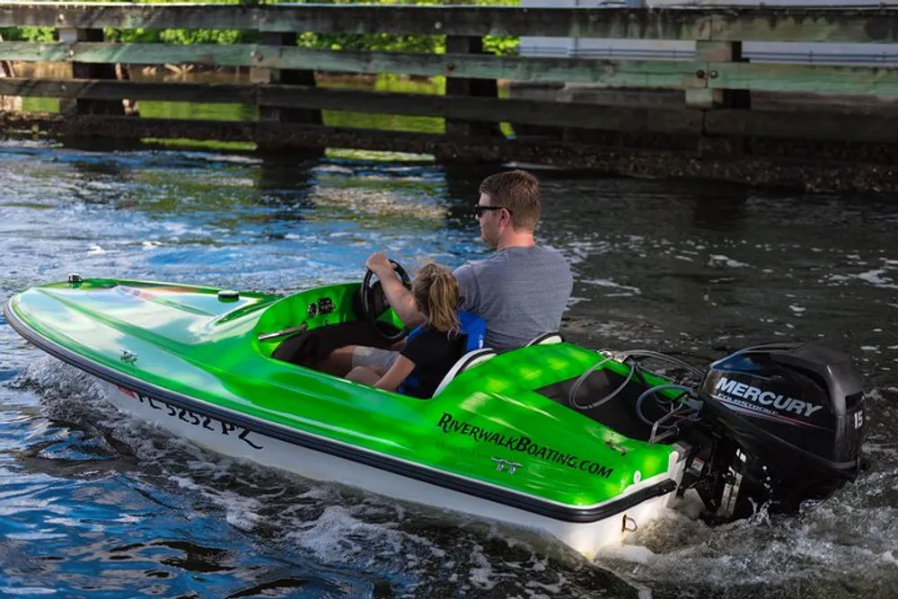 Two people are riding in a bright green and white motorboat as it moves under a wooden bridge